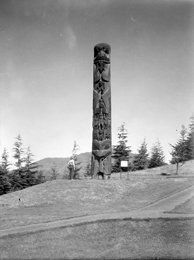 Black and white photograph of a single totem pole on a gentle hill surrounded by well trimmed grass, with a woman standing next to it, looking up