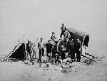 Black and white photograph of a prairie landscape, with a group of people standing in front of a large cart and tent