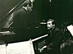 Photograph of Glenn Gould seated at the piano and talking to Walter Susskind, December 1960