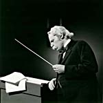 Photograph of Glenn Gould, as Sir Nigel Twitt-Thornwaite, at a lectern with score and baton, 1980